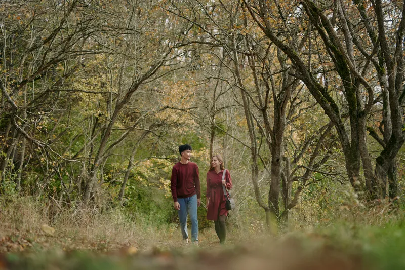 A candid scene of a couple walking along an autumn woodland path, capturing authenticity and warmth in their natural interaction, relaxed posture, genuine shared moment, muted tones and seasonal