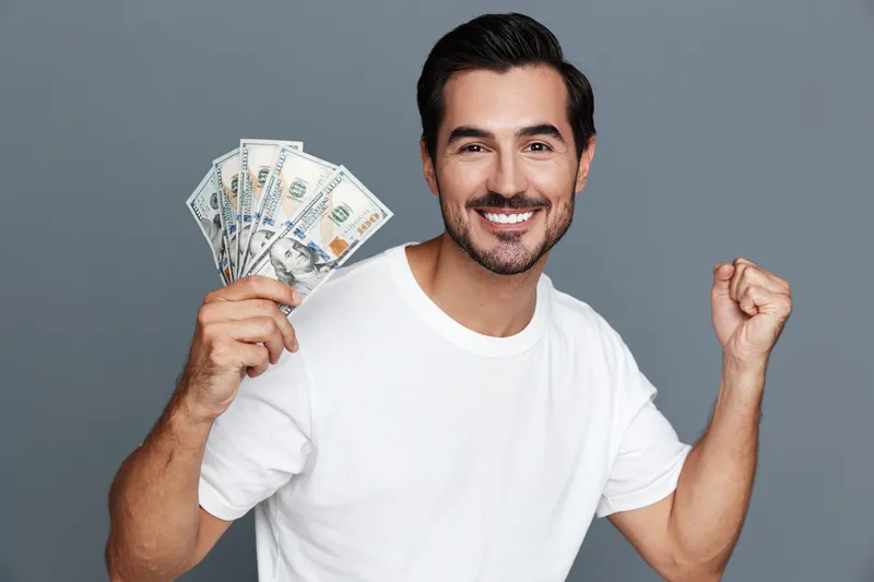 Cheerful young caucasian man holding cash and celebrating success with a big smile, isolated on solid gray background. Financial achievement concept.
