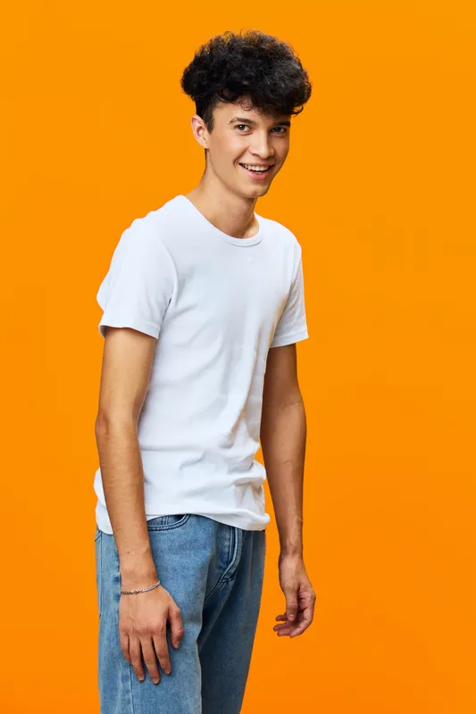 Smiling young man wearing white t-shirt and blue jeans stands against bright orange background with casual posture and friendly expression.