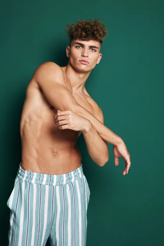 Young man with curly hair posing shirtless against green background wearing striped pants, showing toned muscles and confident expression in studio portrait photography.