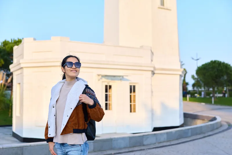 Fashionable woman wearing a colorblocked jacket and oversized sunglasses stands outdoors near a white architectural building, radiating confidence and style under a bright sunny sky.