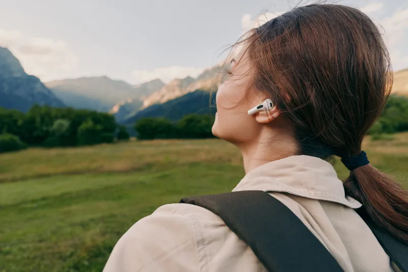 A young woman with a backpack listens to music in earphones while turning her head toward distant mountains in a bright open field, capturing a moment of exploration and calm curiosity.