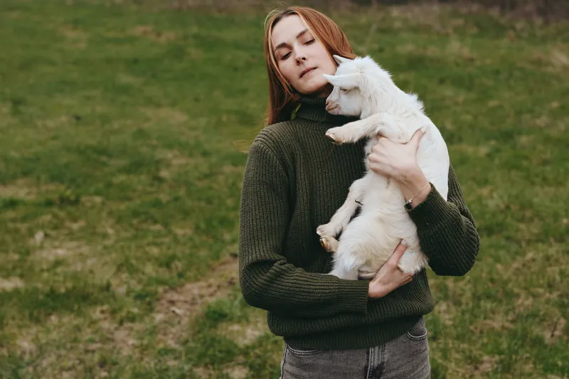 A woman in a dark green sweater cradles a white goat kid in a grassy field, capturing gentle countryside companionship, rural lifestyle, and calm outdoor moments, reflecting connection between