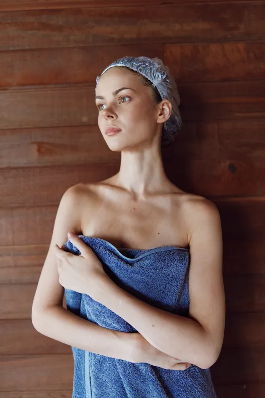 young woman wrapped in towel with shower cap looks thoughtfully wooden background soft natural light calm expression wellness and self-care concept