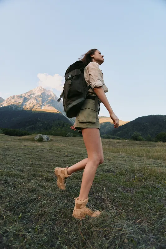 A woman hiking outdoors with a backpack in a mountainous landscape, wearing shorts and boots. A lifestyle moment of adventure, exploration, and outdoor fitness in nature.