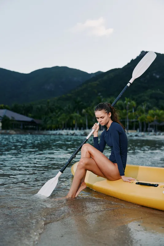 Young woman sitting on yellow kayak in water, holding paddle, wearing navy swimwear near tropical shore with mountains in background.