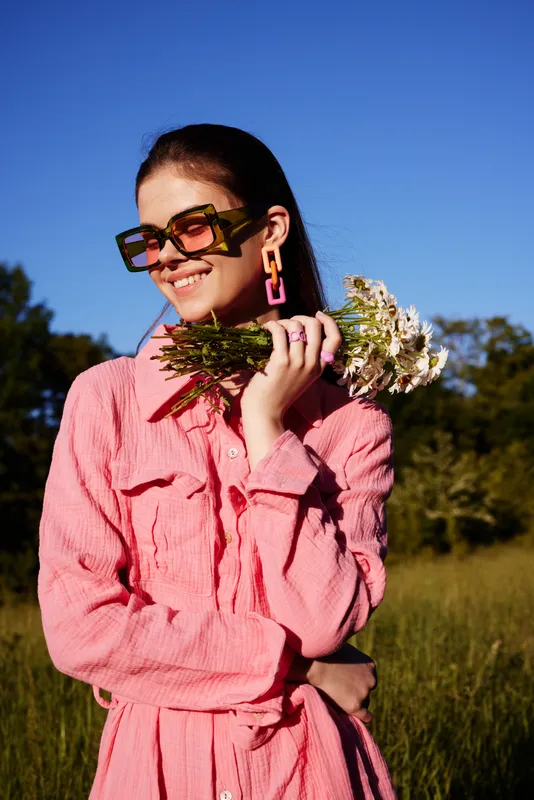 Young woman smiling in a pink dress, holding flowers against a bright blue sky, showcasing summer fashion and vibrant emotions, perfect for seasonal themes