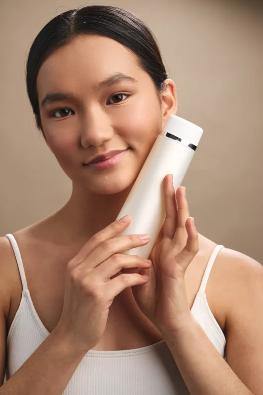 asian woman holds a white bottle close to her face in a calm studio, presenting a skincare product with a gentle smile and confident poise.