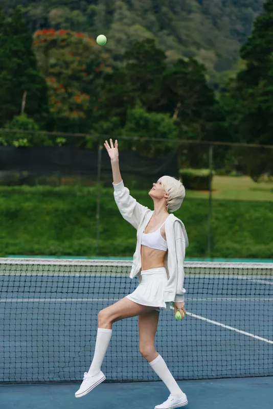 Young female tennis player serving on a court, showcasing athleticism and style in a white outfit, with a vibrant natural background