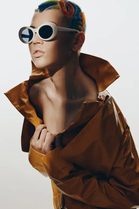 Stylish woman with colorful short hair and white sunglasses, wearing a brown leather jacket with large collar, showing confident and edgy modern fashion look.
