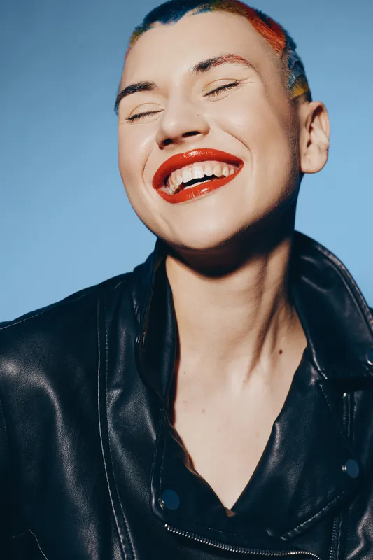 joyful woman with bright red lipstick and colorful hair wearing a black leather jacket against a clean blue background, expressing happiness and confidence.