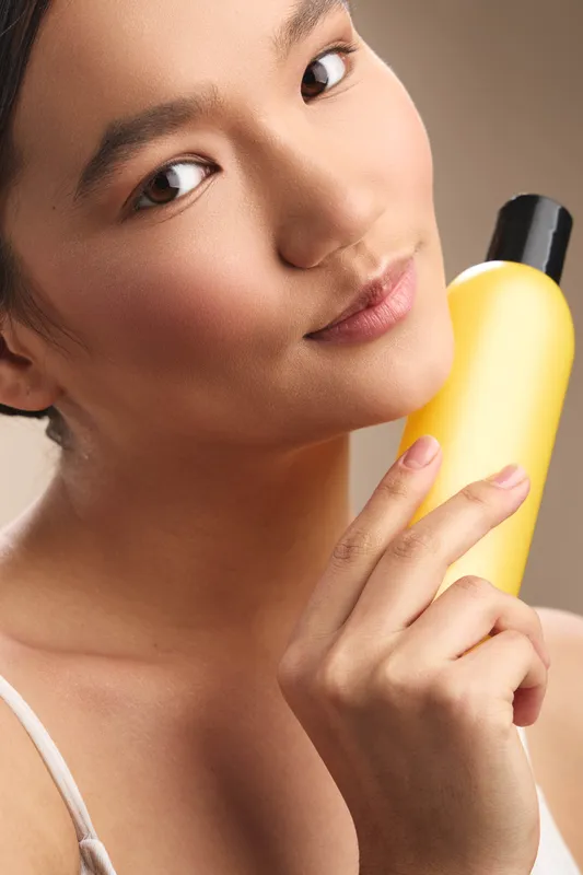 Asian woman holds a bright yellow bottle near her face, presenting skincare product with a gentle smile. The warm lighting highlights smooth skin and a confident, serene mood.