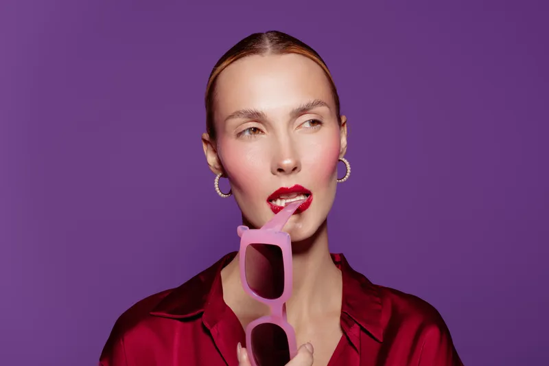 Portrait of a fashionable woman with red lipstick holding pink sunglasses near her lips against purple background. Concept of style, beauty, fashion, and confident attitude.