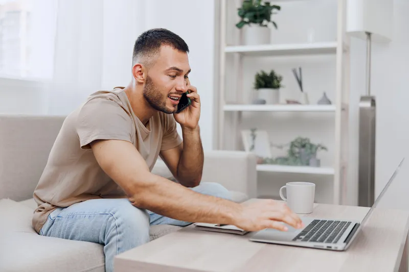 Engaged man making a phone call while working on his laptop at home, showcasing a modern, productive lifestyle in a cozy, contemporary environment.