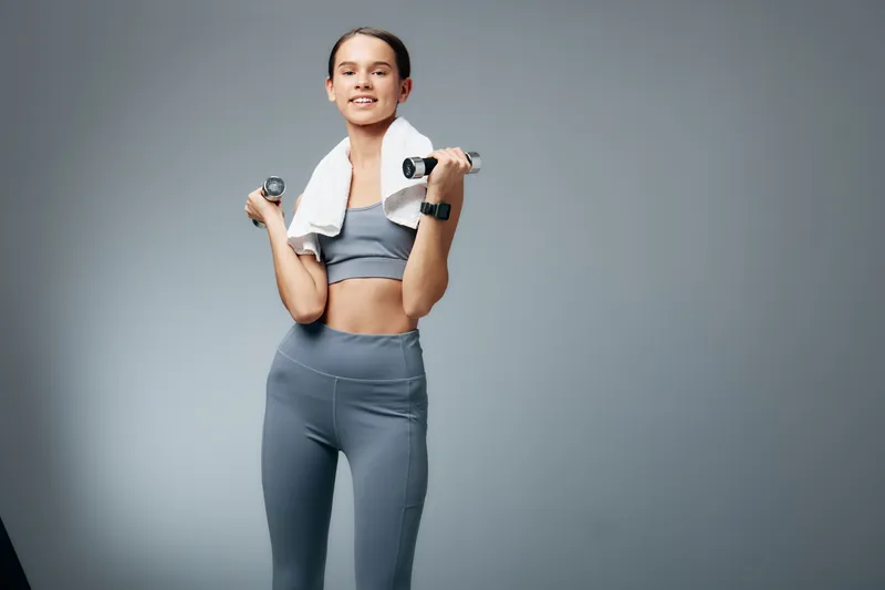 Energetic woman fitness enthusiast lifts dumbbells in a studio setting, wearing athletic wear with a towel over her shoulders, showcasing sport style, strength and active training.