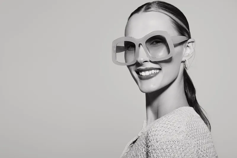 Smiling caucasian woman with pale skin wearing oversized sunglasses and knitted sweater looks happy on neutral background.