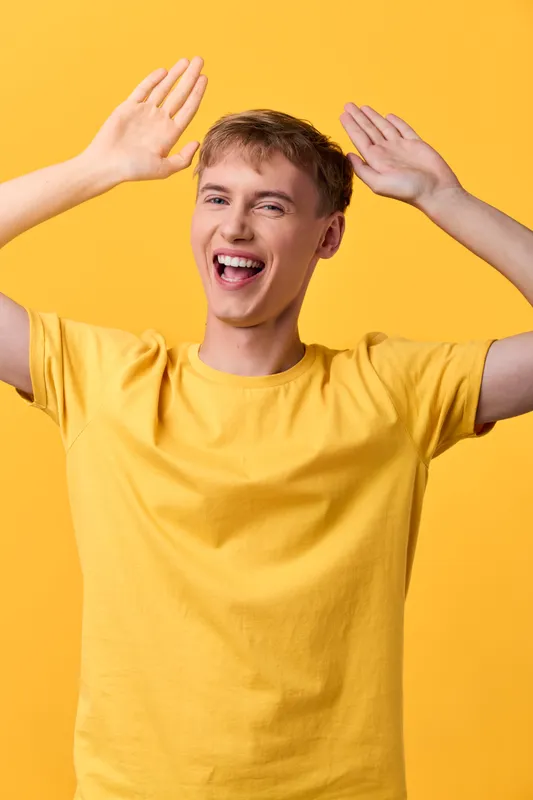 Happy man in a yellow shirt raises arms with a playful smile, bright studio backdrop, conveying energy, positivity, and upbeat mood suitable for lifestyle, advertising, and wellness themes