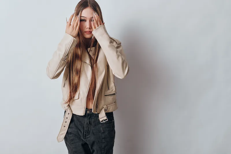 Young woman with long hair wearing beige jacket and black pants posing with hands near face standing against light wall background in casual outfit.
