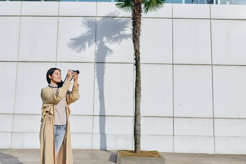 Photography of a woman in a beige coat using a handheld camera beside a palm tree and a clean white wall, conveying urban leisure, travel mood and candid creativity