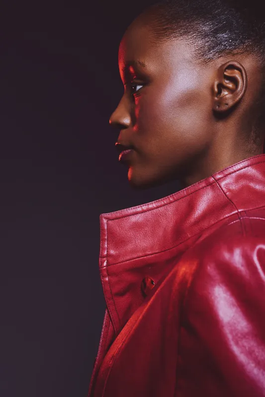 Profile of young African woman with dark skin wearing red leather jacket against black background. Concept for fashion, beauty, confidence, and style with studio lighting and vibrant colors.