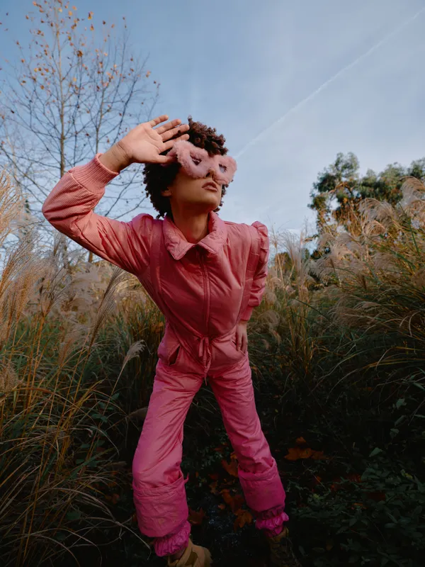 Fashion model in pink jumpsuit posing outdoors with a fluffy mask among tall grasses under a clear sky, artistic portrait capturing autumn mood, bold styling and creative attitude for editorial use