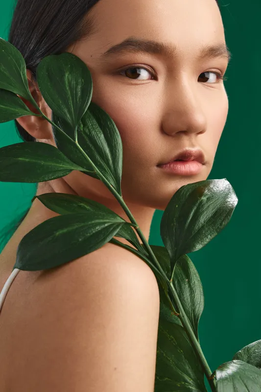 Asian woman photographed in a closeup with a lush leaf against a green backdrop, conveying calm beauty, natural textures, and a modern minimal studio mood.