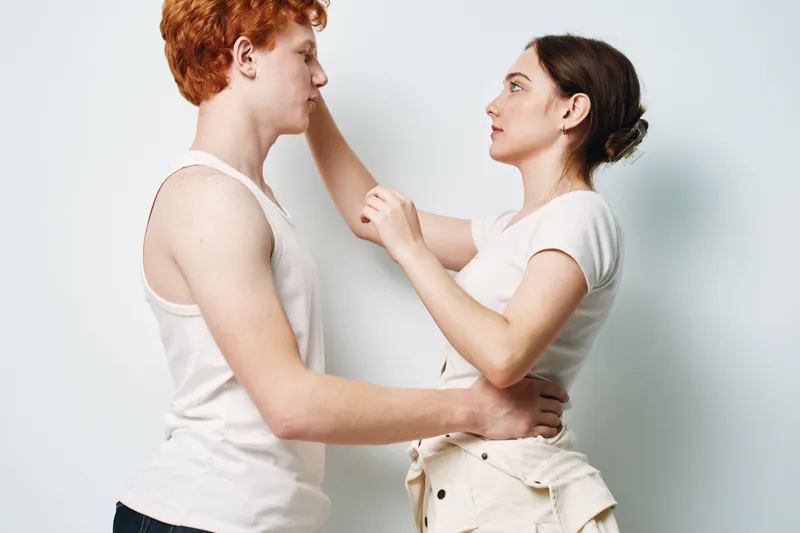 Young couple standing face to face in white casual clothes, woman touching man’s face, intimacy and connection in light studio background.
