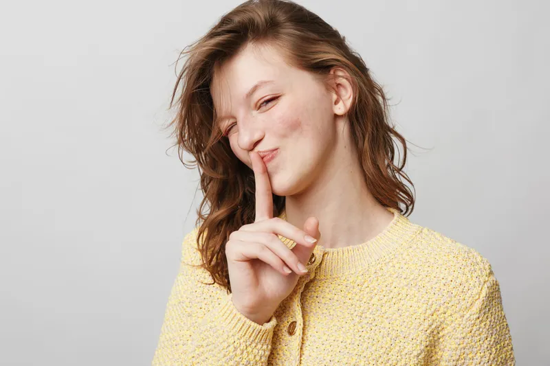 Young woman in yellow sweater makes shushing gesture with finger on lips, smiling slightly. Studio portrait on grey background. Concept of silence, secret, and quiet communication.