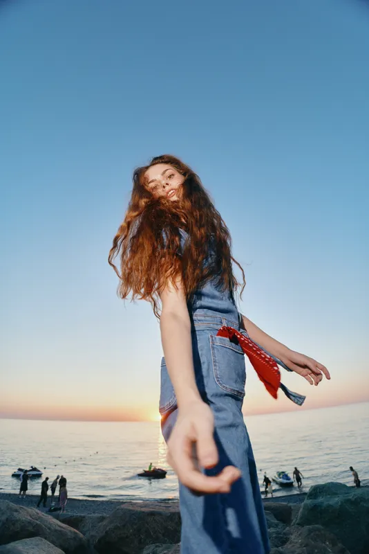 Lifestyle woman with modern boho-western denim street style and flowing hair, captured in warm film color tones by the ocean at sunset, radiating natural freedom and youthfulness.