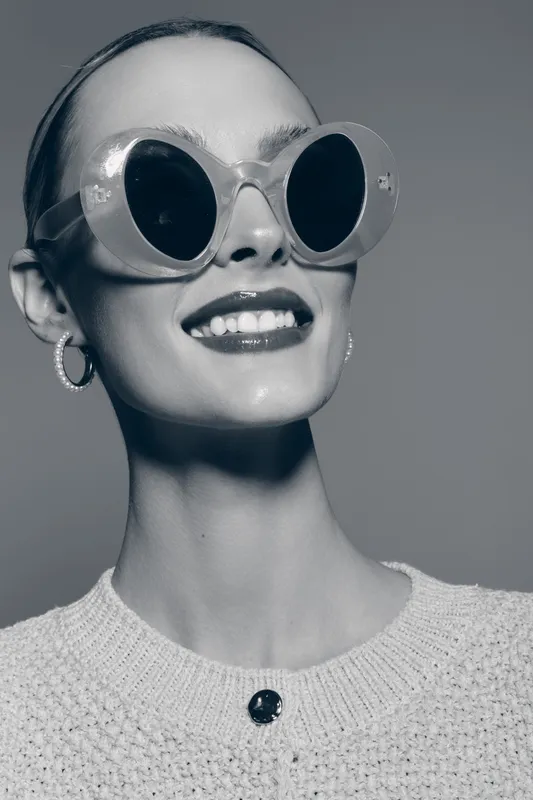 Smiling slim white woman with pale skin wearing large round sunglasses and hoop earrings. Studio portrait with knitted sweater on grey background shows fashion and confidence.