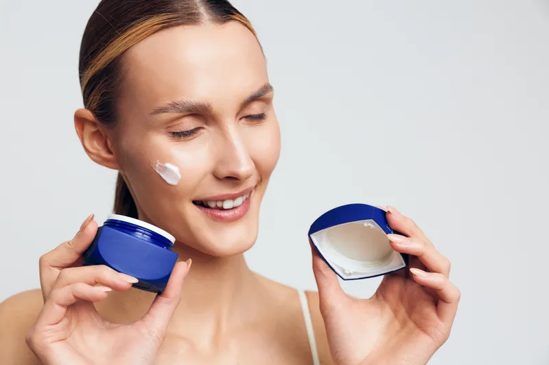 Young woman with blonde hair applies moisturizing cream on her cheek while holding an open blue jar and smiling with closed eyes against a white background.