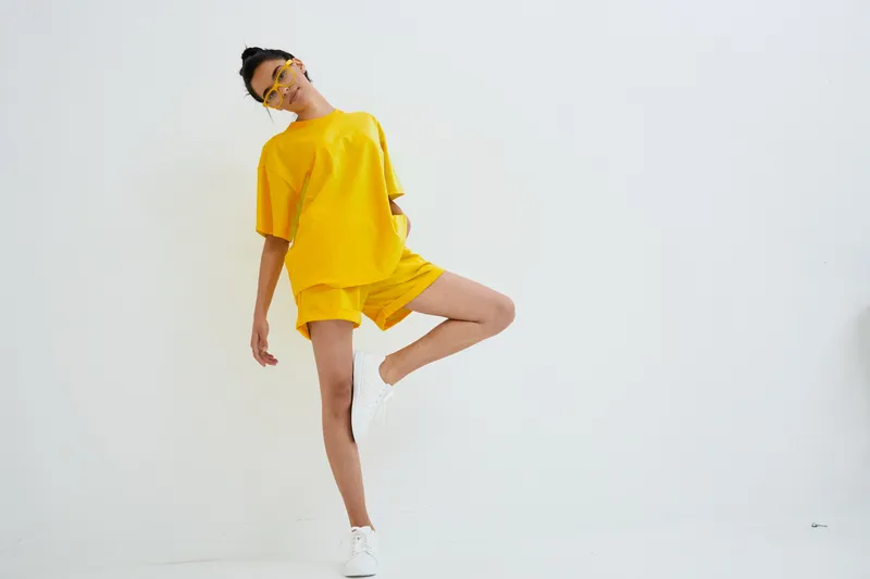 Young woman in yellow casual outfit posing playfully against white wall, wearing white sneakers and sunglasses, showcasing modern street style and vibrant summer vibe.