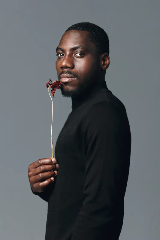 African man with dark skin wearing black turtleneck holding dried flower in creative conceptual visual style studio portrait with neutral gray background and serious expression.