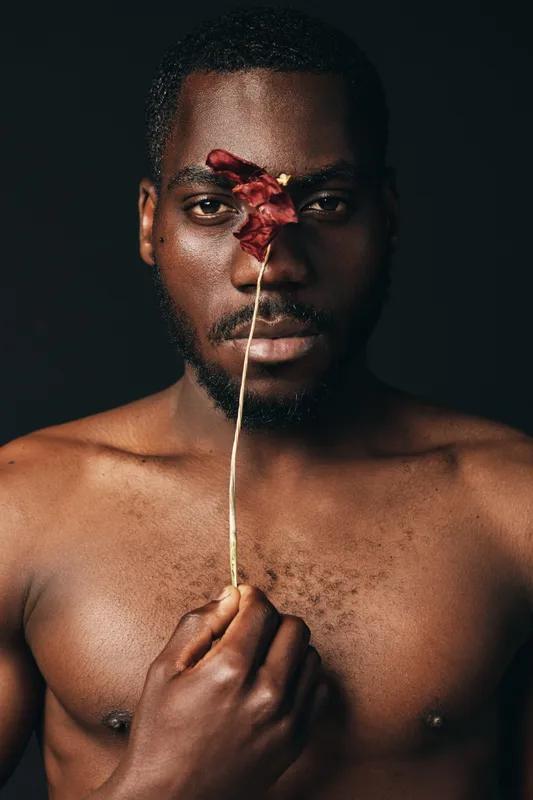 Black man with dark skin holding red flower in front of face in creative visual design style. Portrait with bare chest and artistic expression on dark background.