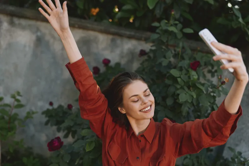 Young woman lifestyle enthusiast celebrates with arms raised, smiling, taking selfie outdoors among roses and a garden wall, wearing a red shirt in sunny daylight, bright candid moment
