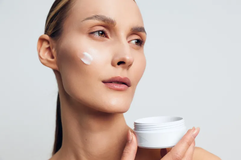 Young woman applying face cream, close up portrait, natural skin care routine, minimal makeup, soft lighting, neutral background, healthy glowing complexion