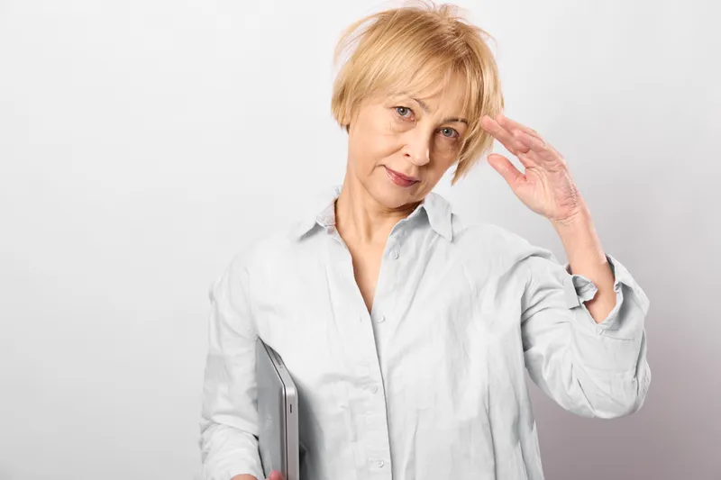 Woman senior smartphone portrait white indoors hand gesture blonde casual, studio lighting, calm expression, light touch near face, holding device, soft minimal background