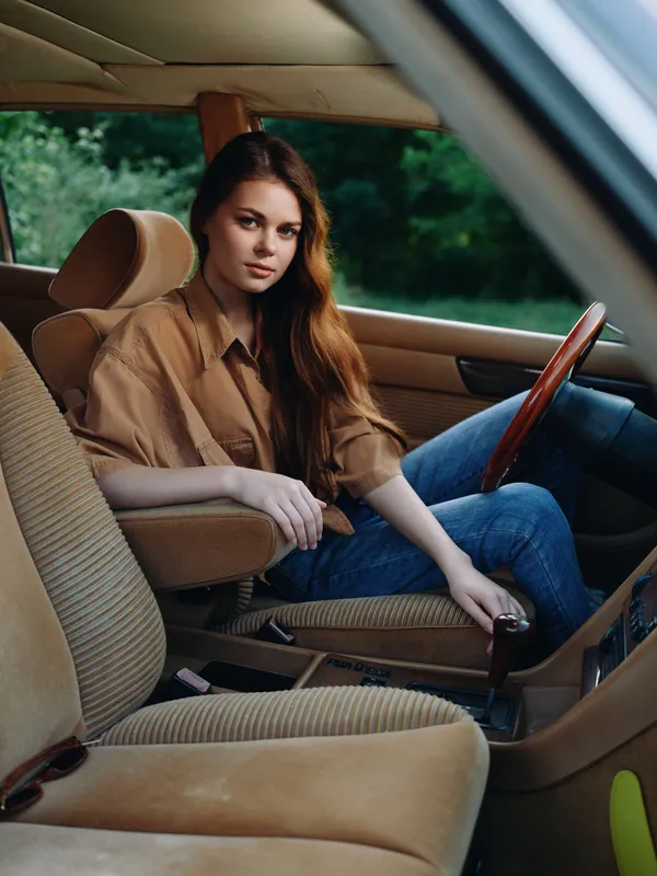 Young woman in stylish casual outfit sitting in a vintage car, exuding confidence and relaxation amidst a natural green background.