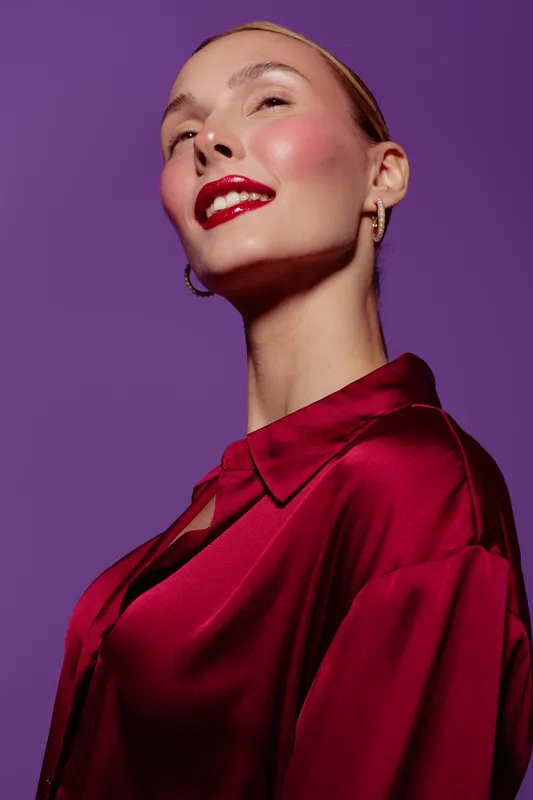 Confident woman with red lipstick and blush wears shiny red blouse against purple background. Portrait with elegant makeup and stylish earrings. Concept of beauty, fashion, and glamour.