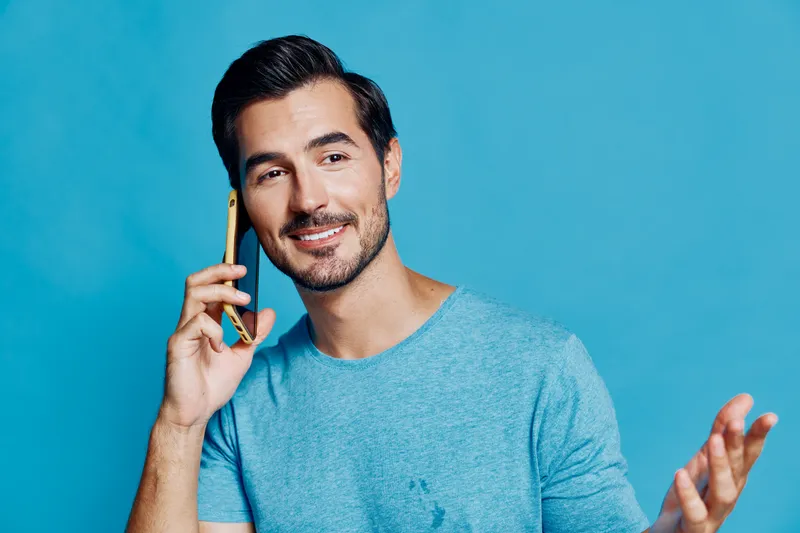 Smiling young cheerful man with dark hair wearing blue t-shirt talking on mobile phone, looking at the camera, isolated on solid blue background. People lifestyle concept.