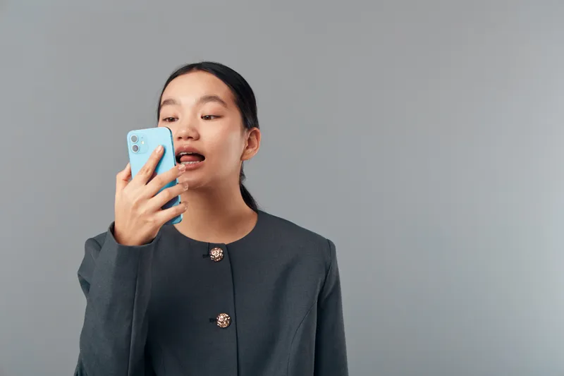 Young woman speaking into smartphone for voice message or call with focused expression wearing dark jacket on gray background. Female person using mobile phone for communication or technology.