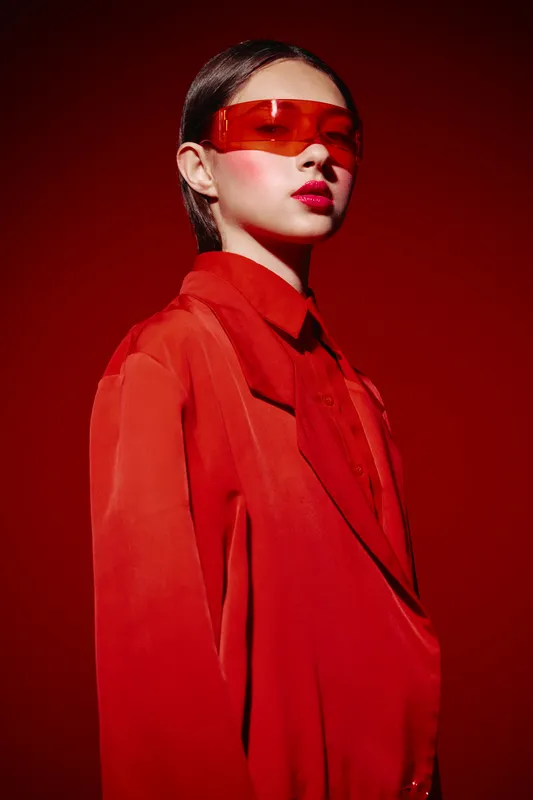 Young woman in oversized red blazer and matching red tinted sunglasses poses confidently against red background. The fashion creative concept featuring bold color and attitude.