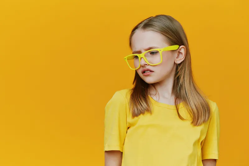 A young girl with long hair wearing yellow glasses and a yellow t shirt against a bright yellow background, expressing a playful emotion and youthful spirit