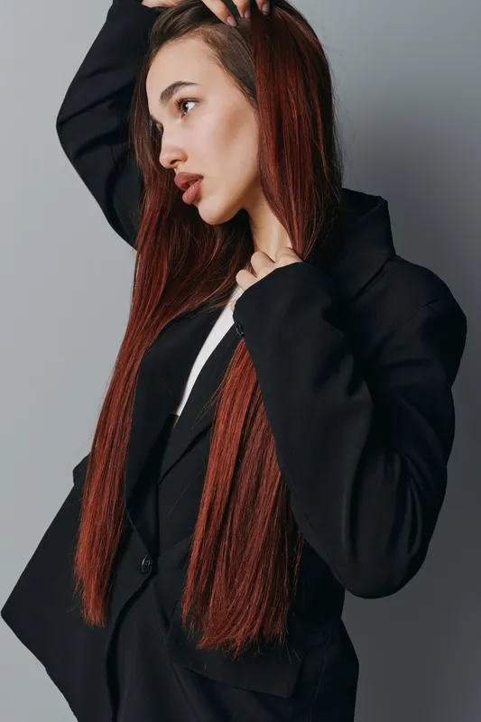 Woman with long straight hair wearing black blazer posing in studio against gray background. Female model with natural makeup and serious expression in fashion portrait style.