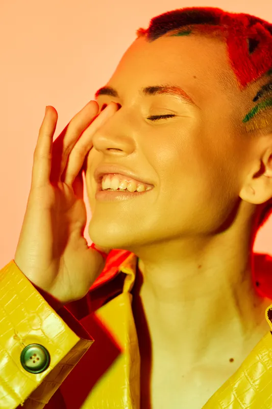 smiling woman with colorful red hair and yellow jacket enjoying bright studio light, closed eyes, joyful expression, vibrant warm tones, close-up portrait