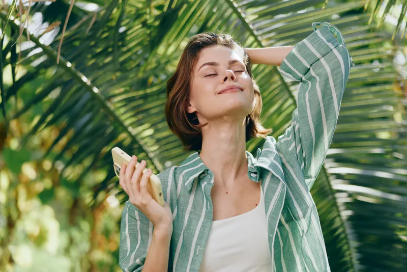 A cheerful woman stands outdoors among lush palm leaves, smiling with eyes closed, wearing a light striped shirt and holding a small object.