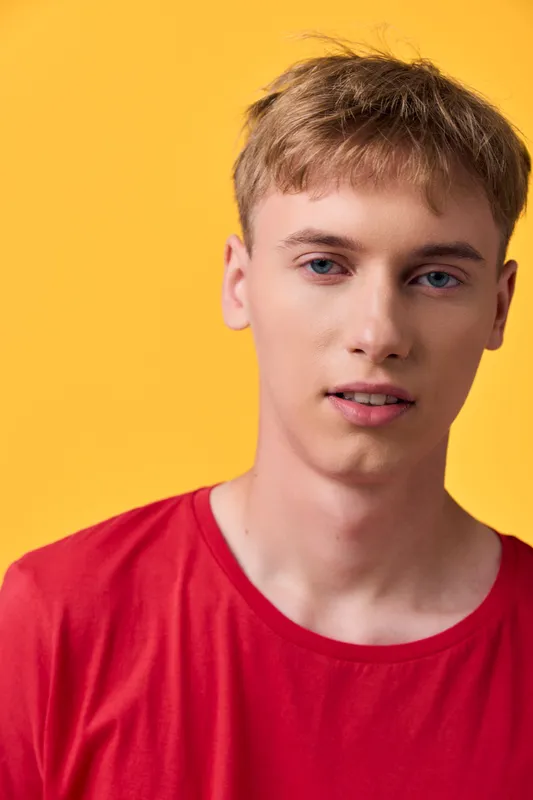 Young man in a red shirt stands against a bright yellow backdrop, creating a bold studio style. Clean lighting highlights features and casual mood, ideal for lifestyle, fashion, product, or creative