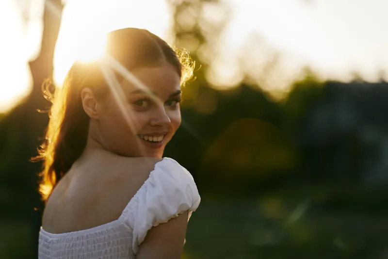 Cheerful young woman in white dress enjoying sunset, smiling, with natural green background and warm sunlight creating a joyful atmosphere