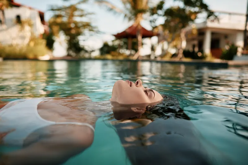 Young woman in white swimsuit relaxes floating on water surface in outdoor pool with blurred tropical background enjoying calm lifestyle and peaceful moment under sunlight.