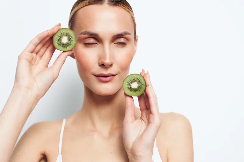 Young woman holding two halves of fresh kiwi fruit near her face with closed eyes, minimal makeup, soft lighting, smooth skin, isolated on white background, healthy lifestyle concept